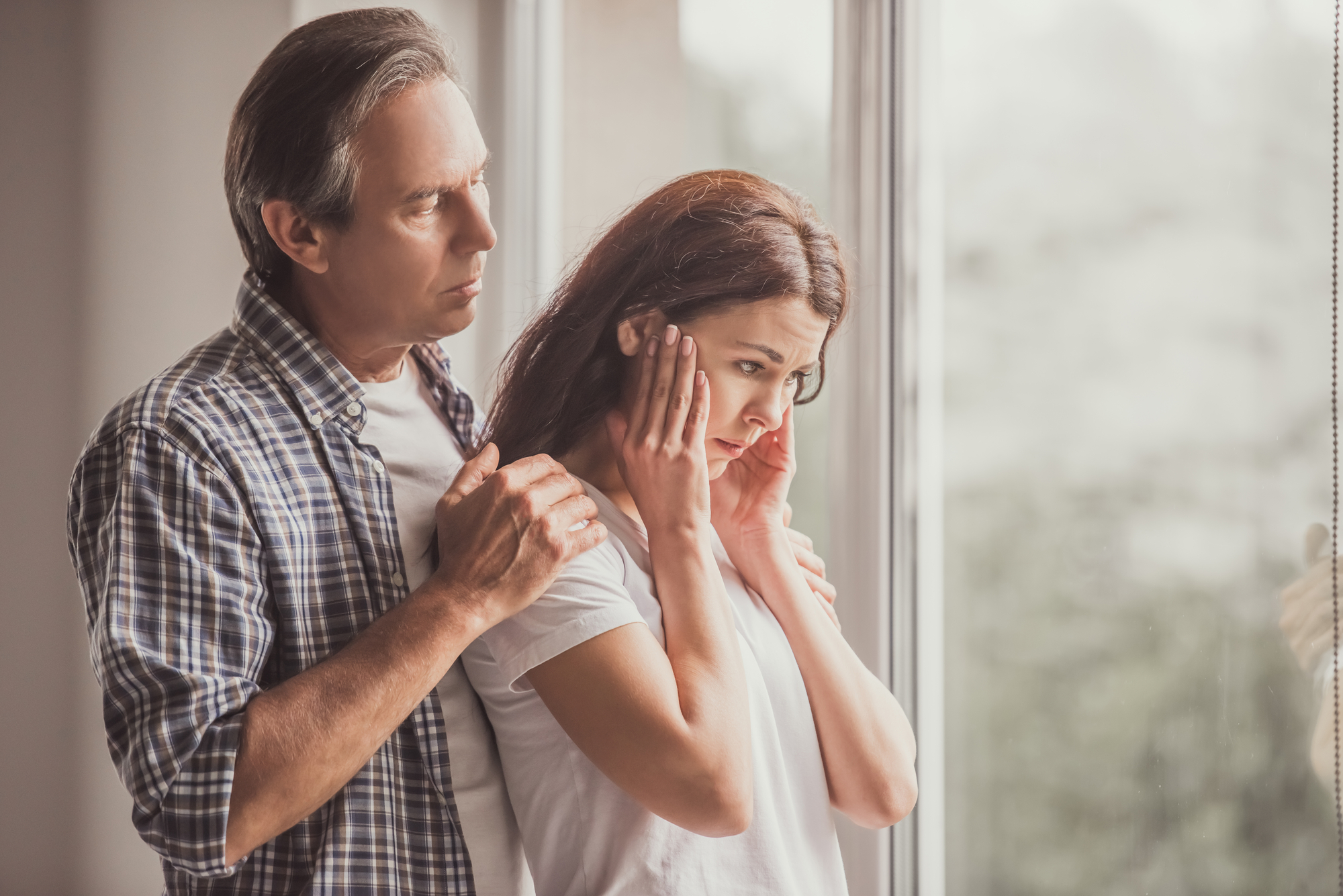 A man stands close to a woman, gently holding her shoulders, looking concerned. The woman faces away, pressing her hands to her temples, appearing distressed or deep in thought. They are indoors, next to a large window with soft daylight filtering in.