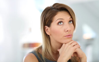 A woman with straight, shoulder-length blonde hair gazes upward with her chin resting on her clasped hands. She has a thoughtful expression and is wearing a sleeveless gray top. The background is blurred and bright, suggesting an indoor setting.