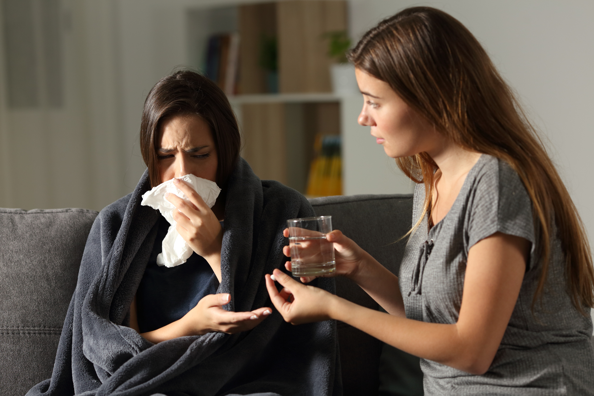 A woman wrapped in a blanket is sitting on a couch, holding a tissue to her nose with one hand and her forehead with the other, appearing unwell. Another woman is sitting next to her, offering a glass of water and looking concerned.