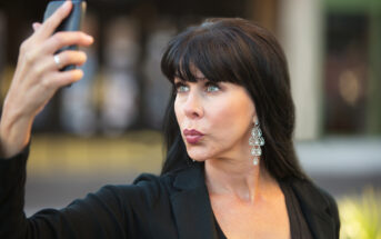 A woman with long dark hair wearing a black outfit takes a selfie outdoors. She is posing with her lips pursed and eyes looking upwards. She is wearing large dangling earrings, and there is a blurred background with hints of greenery and a building.