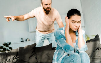 A man stands behind a woman, angrily pointing and shouting. The woman, sitting on a couch, covers her ears with her hands, looking distressed. The scene takes place in a brightly lit living room with modern decor.