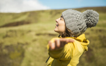 A person wearing a gray knit hat with a pom-pom and a yellow jacket stands outdoors with their eyes closed and arms outstretched, enjoying the sun. They are in a grassy landscape with a blurred background of hills and a partly cloudy sky.