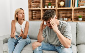 A woman and a man sitting on a gray couch appear to be in a heated discussion. The woman, with a frustrated expression, gestures with her hands, while the man holds his head in his hands, looking distressed. A wooden shelf with books and plants is in the background.