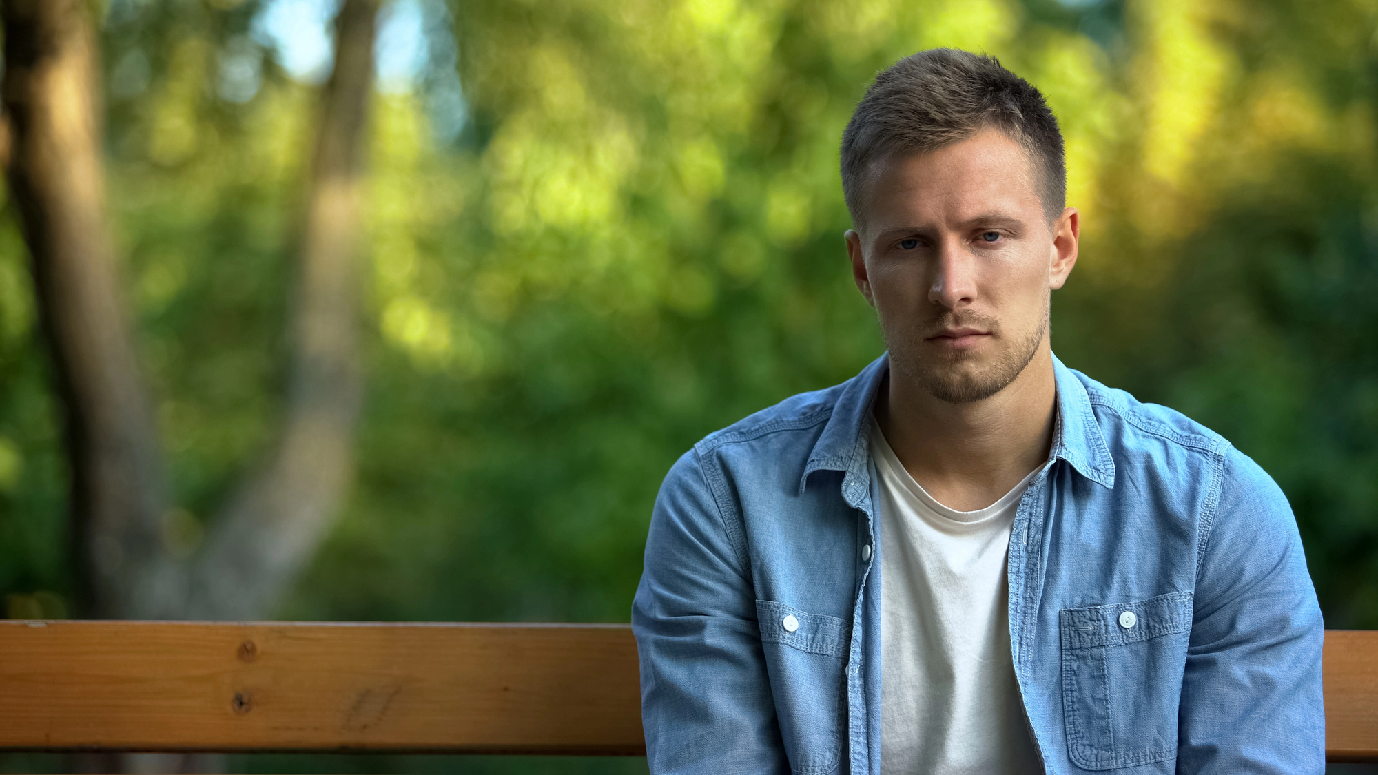 A man with short hair and a trimmed beard is sitting on an outdoor wooden bench. He is wearing a light blue denim shirt over a white T-shirt. The background features blurred greenery, suggesting a park or natural setting. His expression appears thoughtful or somber.