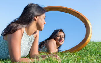 A woman with long dark hair is laying on green grass, facing a large oval mirror that rests on the ground. She is wearing a light green sleeveless top and smiling at her reflection. The sky is bright blue and clear.