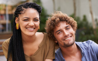 A smiling woman with long braided hair and yellow earrings sits next to a smiling man with wavy hair and a beard. They are both casually dressed and are outdoors with blurred greenery in the background.