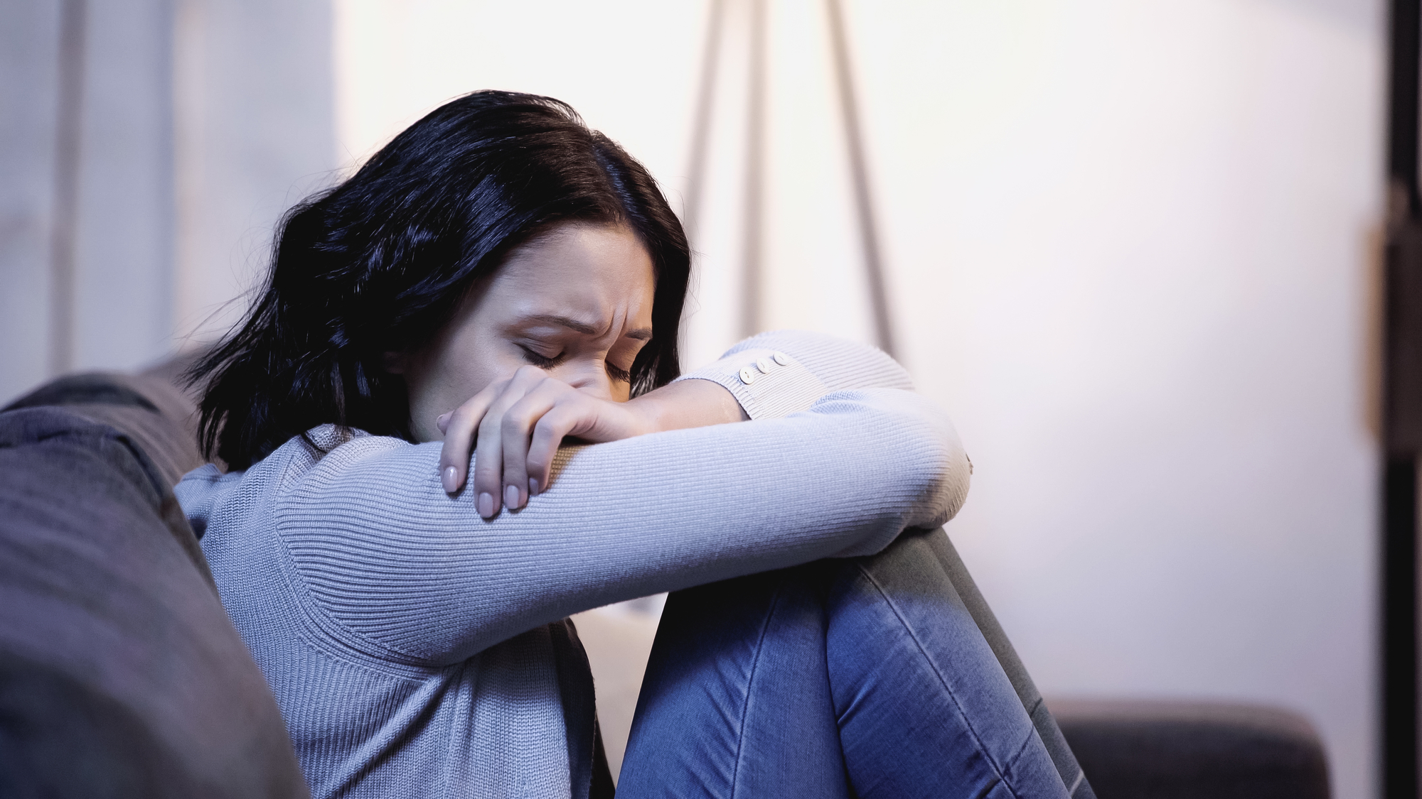 A woman with dark hair sits on a couch with her knees drawn up, hugging her knees and resting her head on her arms. She appears to be upset or in distress, wearing a light gray sweater and jeans. The background is softly lit.