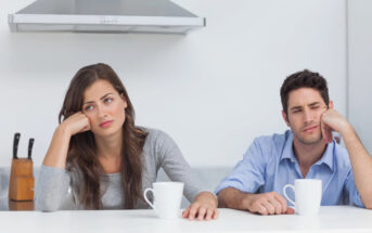 A woman and a man sit at a white kitchen counter with white mugs in front of them. Both appear to be bored or exhausted, resting their heads on their hands. A knife block and a stainless steel range hood are visible in the background.