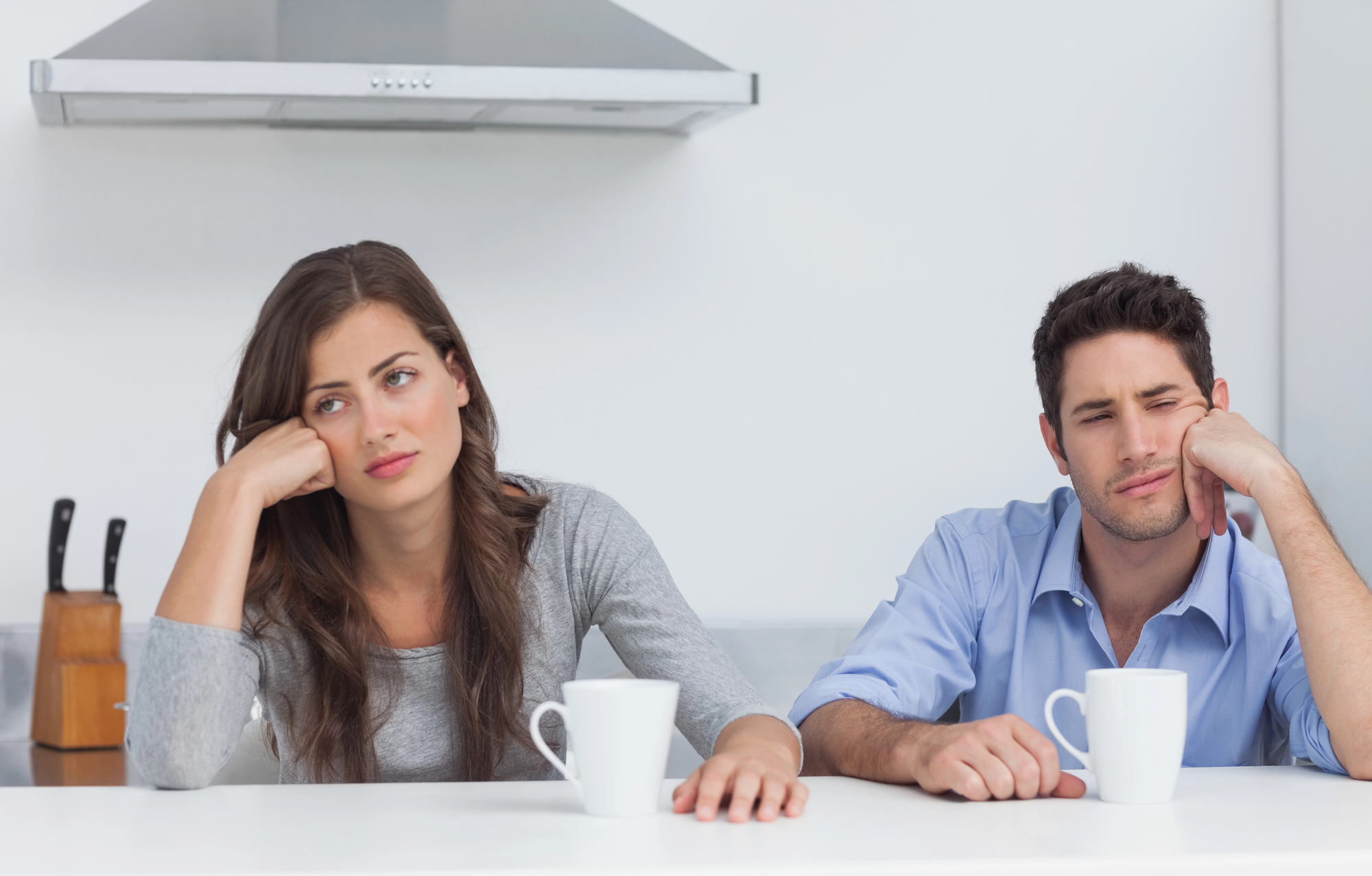 A woman and a man sit at a white kitchen counter with white mugs in front of them. Both appear to be bored or exhausted, resting their heads on their hands. A knife block and a stainless steel range hood are visible in the background.