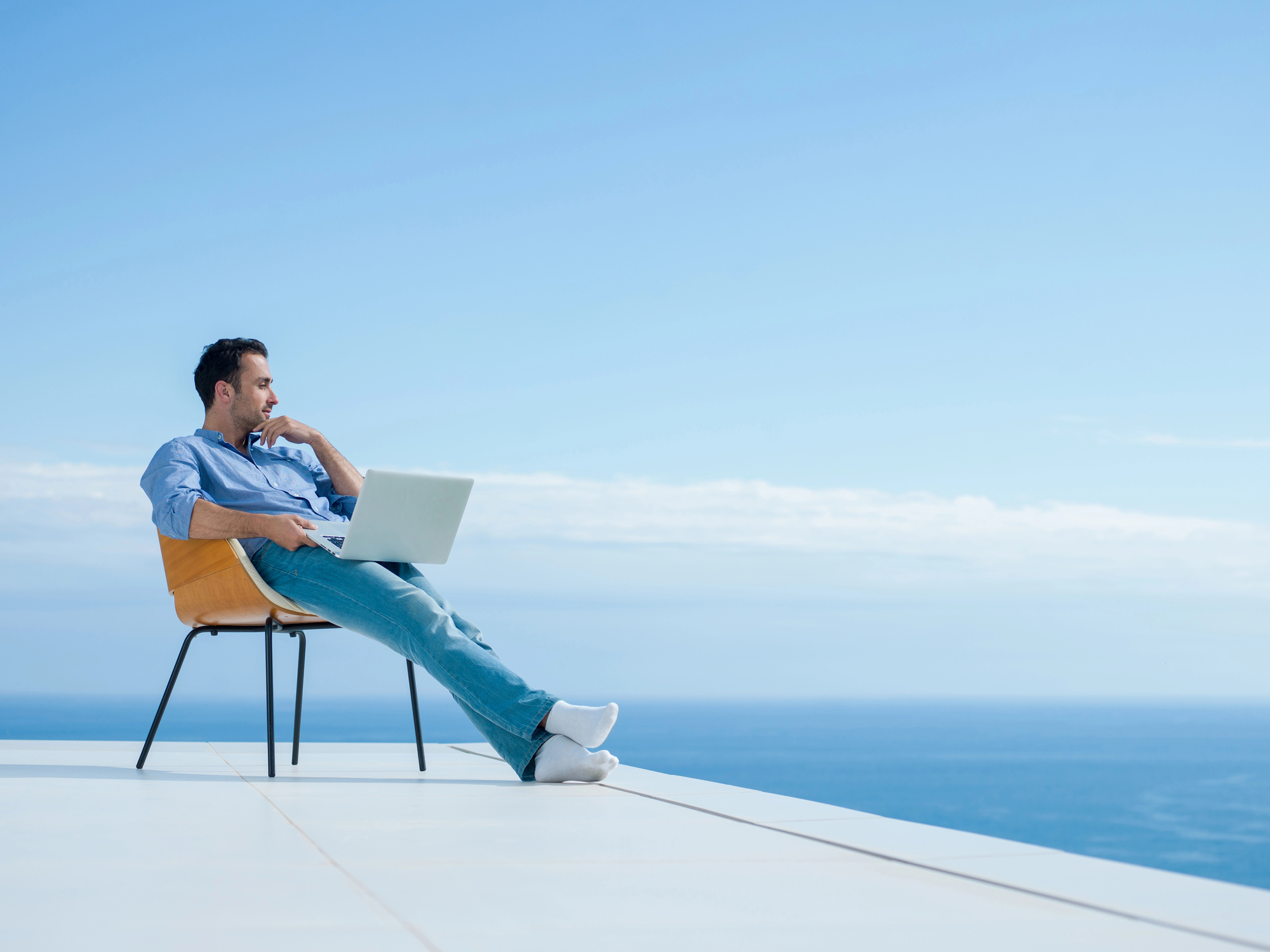 A man sits on an orange chair on a white terrace overlooking a vast, calm ocean under a clear blue sky. He rests his legs on the terrace edge, working on a laptop placed on his lap, one hand on his chin in a thoughtful pose.