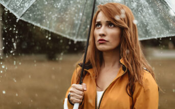 A woman with long brown hair stands outdoors under a clear umbrella, wearing a yellow raincoat. She looks upwards with a slightly displeased expression as raindrops fall around her. The background is blurred, showing a natural, possibly park-like setting.