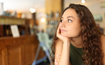 A woman with curly brown hair rests her chin on her hand while looking thoughtfully into the distance. She is seated indoors in what appears to be a cozy cafe with warm lighting and wooden furniture in the background.