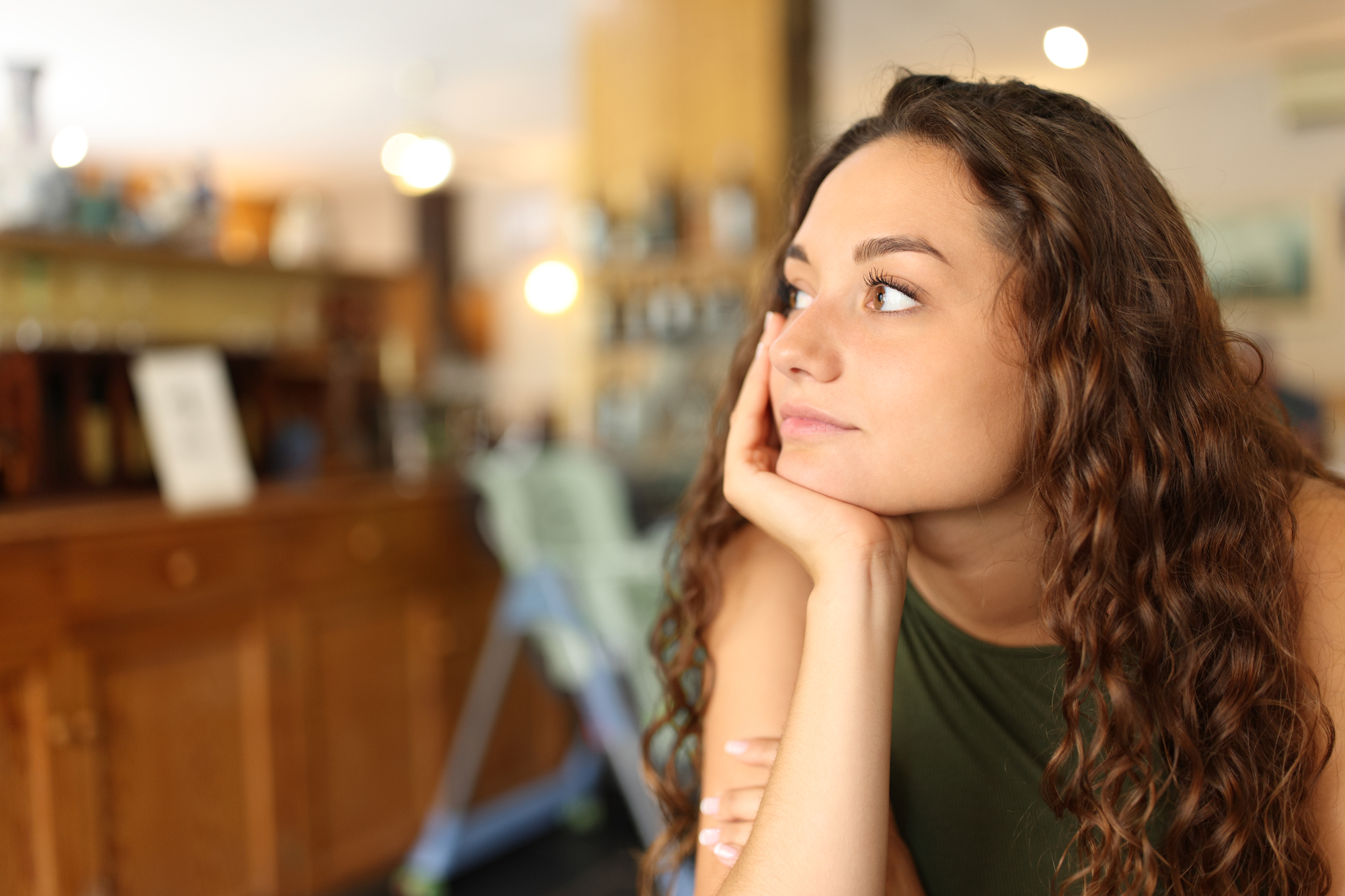 A woman with curly brown hair rests her chin on her hand while looking thoughtfully into the distance. She is seated indoors in what appears to be a cozy cafe with warm lighting and wooden furniture in the background.