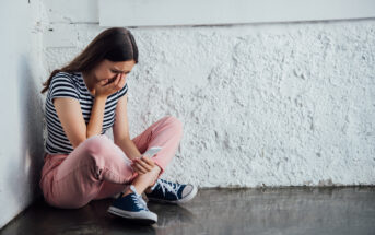 A young woman with long hair, wearing a black and white striped shirt and pink pants, sits on the floor against a white rough wall. She looks distressed, holding a smartphone in one hand and covering part of her face with the other.