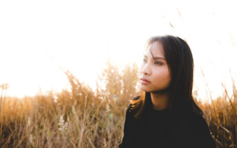 A woman with long black hair wearing a black top looks thoughtfully into the distance while sitting in a grassy field. The sun is shining brightly behind her, creating a warm, golden backlight that illuminates the tall grasses around her.