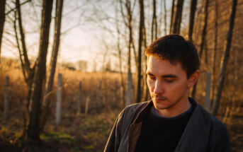 A man stands in a sunlit forest with bare trees, looking thoughtfully downward. The forest floor has patches of green grass, and the background is warmly lit with the glow of the setting sun casting a golden hue. The man is wearing a dark jacket.