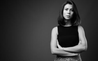 Black and white image of a woman standing with arms crossed against a dark background. She has shoulder-length hair, wears a sleeveless top, and looks seriously at the camera. Her facial expression is composed and confident.