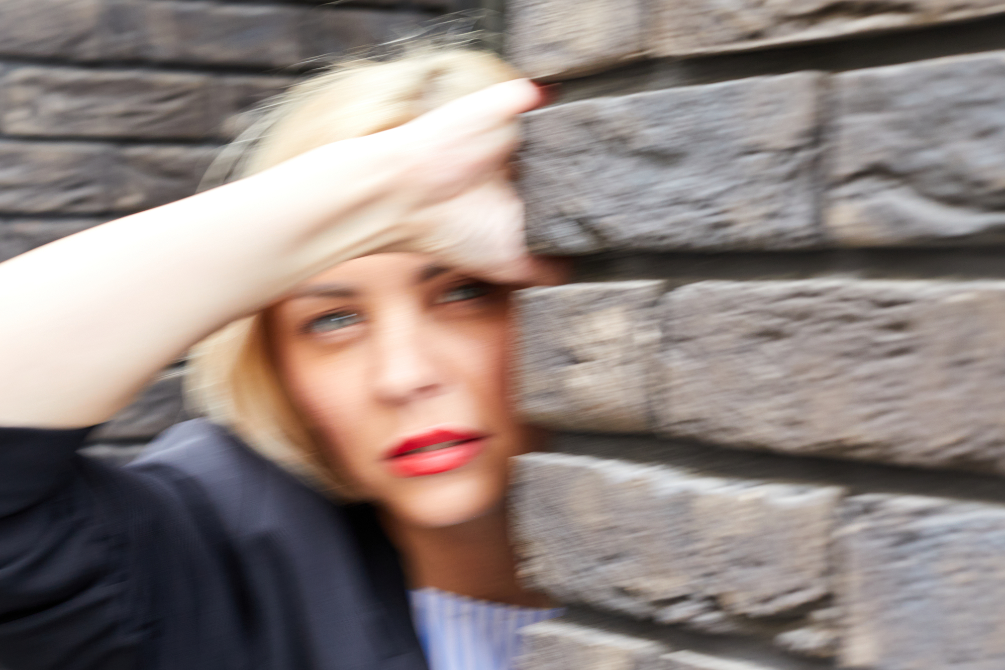 A woman with short blonde hair and red lipstick leans against a brick wall with her arm resting on her forehead. The image is blurred, giving it a slightly dreamy and abstract feel. She is wearing a dark jacket.