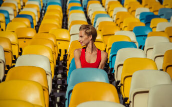 A person with short hair in a red tank top sits among colorful stadium seats, predominantly yellow, blue, and white. The person is smiling and looking to the side, creating a sense of joyful anticipation. The seats around the person are mostly empty.