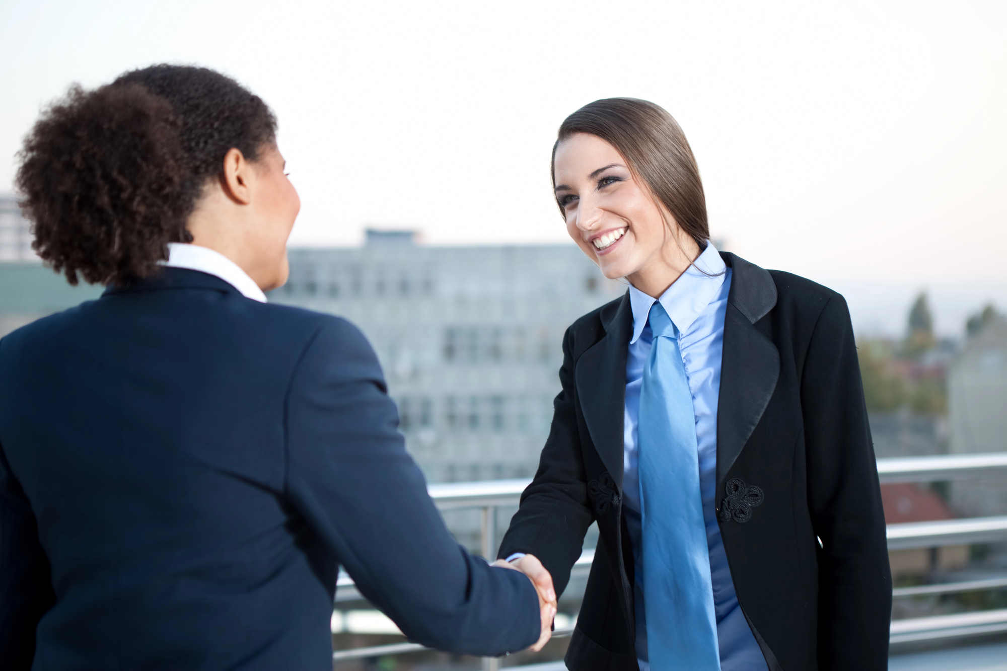 Two businesswomen in suits are shaking hands outdoors on a rooftop with a city view. One has curly hair and the other has straight hair. Both are smiling, indicating a positive interaction or agreement.