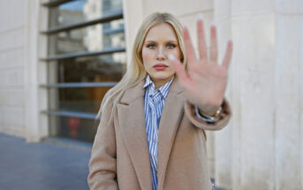 A woman with long blonde hair stands outside an urban building, looking directly at the camera. She is wearing a tan coat over a blue-and-white pinstriped shirt and holds her hand out in front of her, with her palm open and facing the camera.