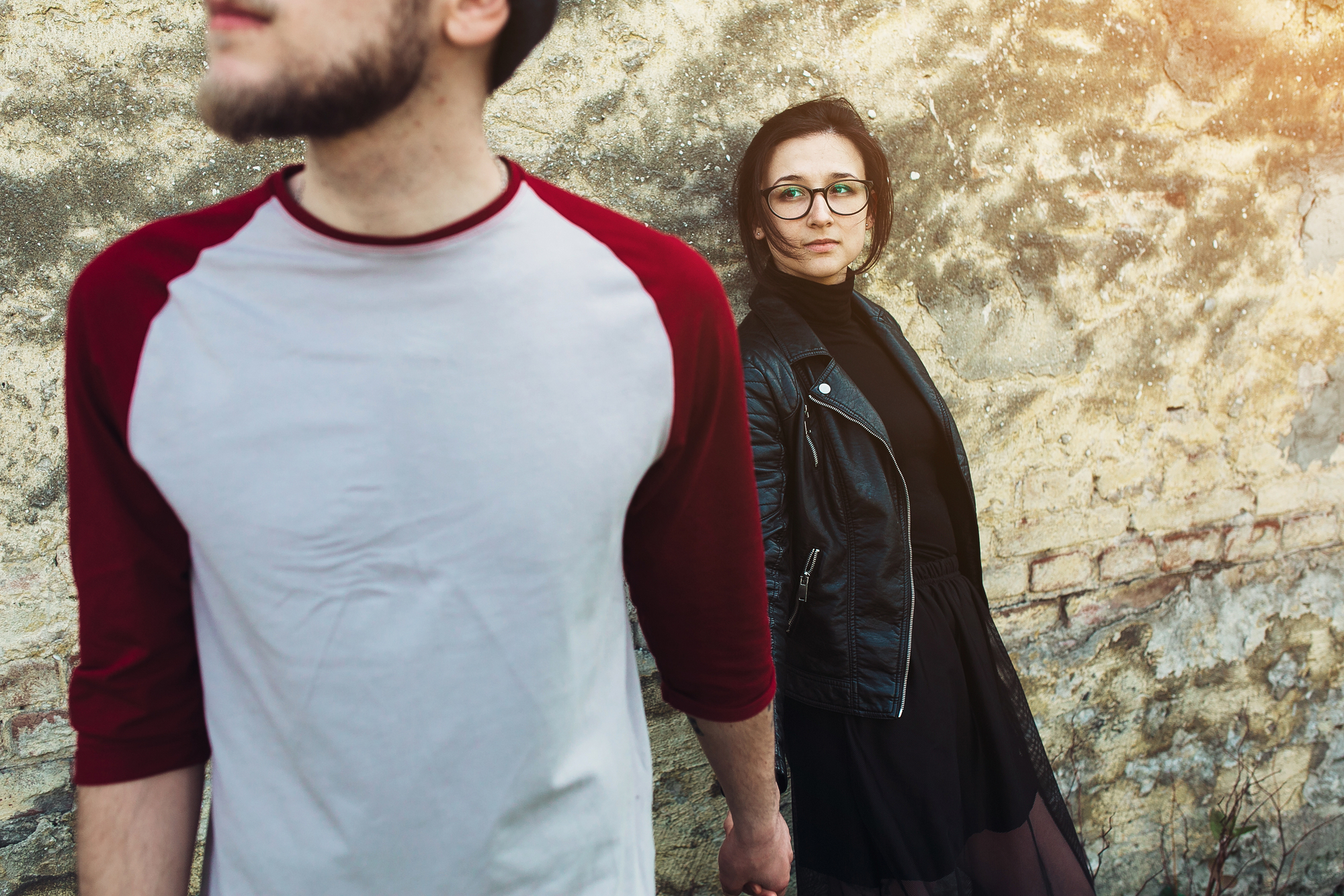 A man and woman stand against a weathered wall. The man, partially out of frame, wears a baseball-style shirt. The woman, in glasses and a leather jacket, looks past the camera. They hold hands, creating a contrast between their disjointed gazes and connected hands.