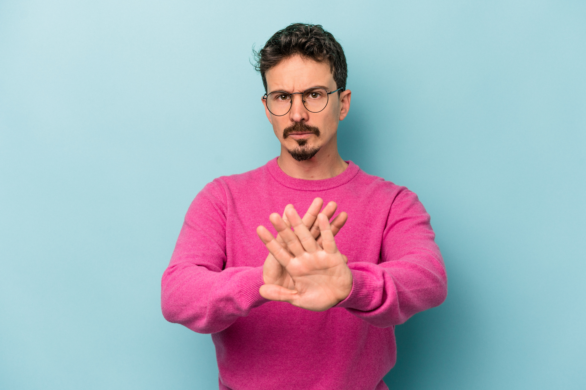 A man with short hair and a goatee, wearing glasses and a pink sweater, stands against a light blue background. He has a serious expression and is showing both palms outward in a stop or halt gesture.