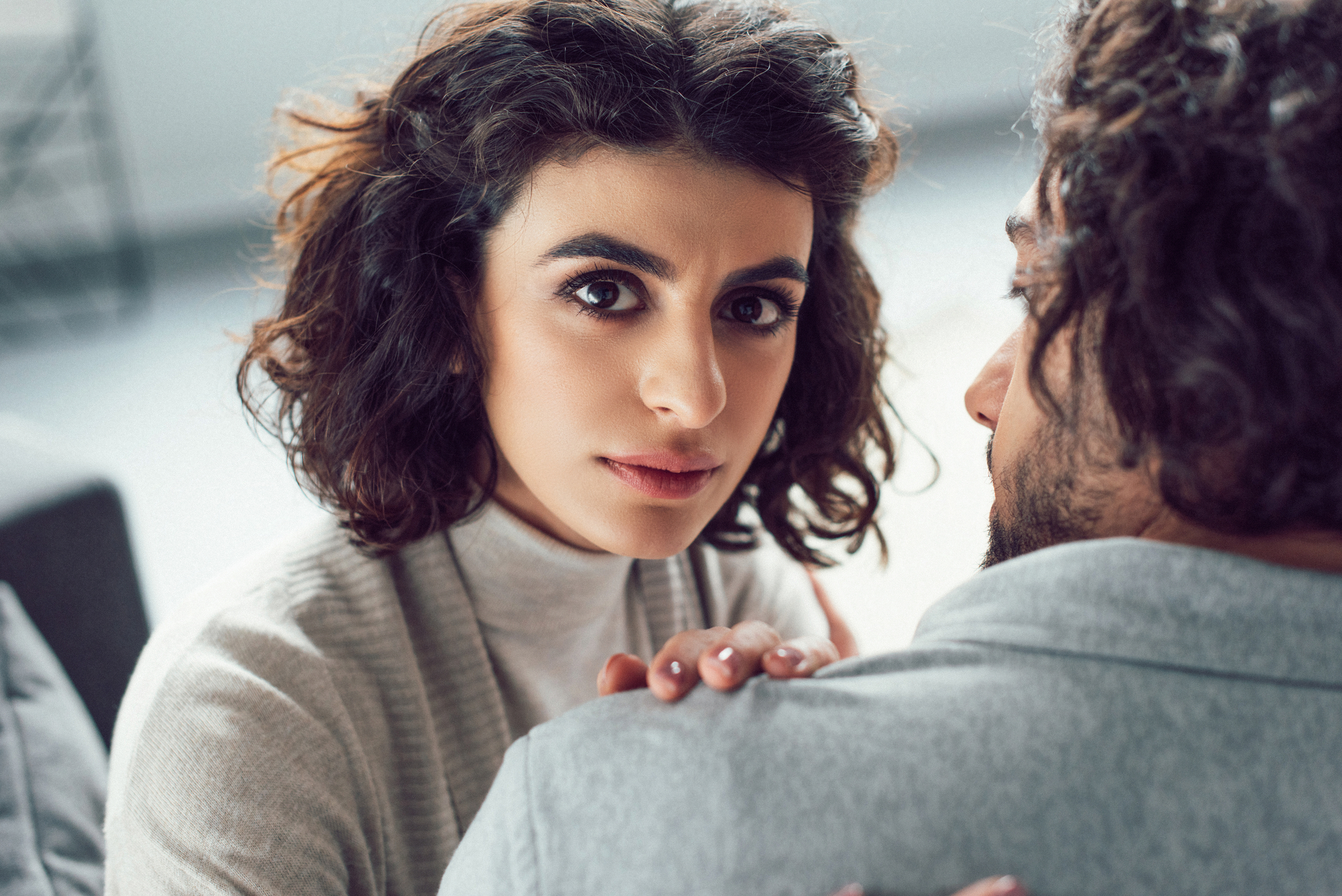 A woman with curly dark hair and expressive eyes looks directly at the camera with a serious expression. She rests her hand on the shoulder of a man with curly hair whose face is partially turned away from the camera. The background is blurred and light-colored.