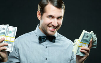 A man wearing a light gray shirt and a black bow tie is holding stacks of US dollar bills in both hands. He has a mischievous smile and is looking slightly to the side. The background is dark, highlighting the man and the money.