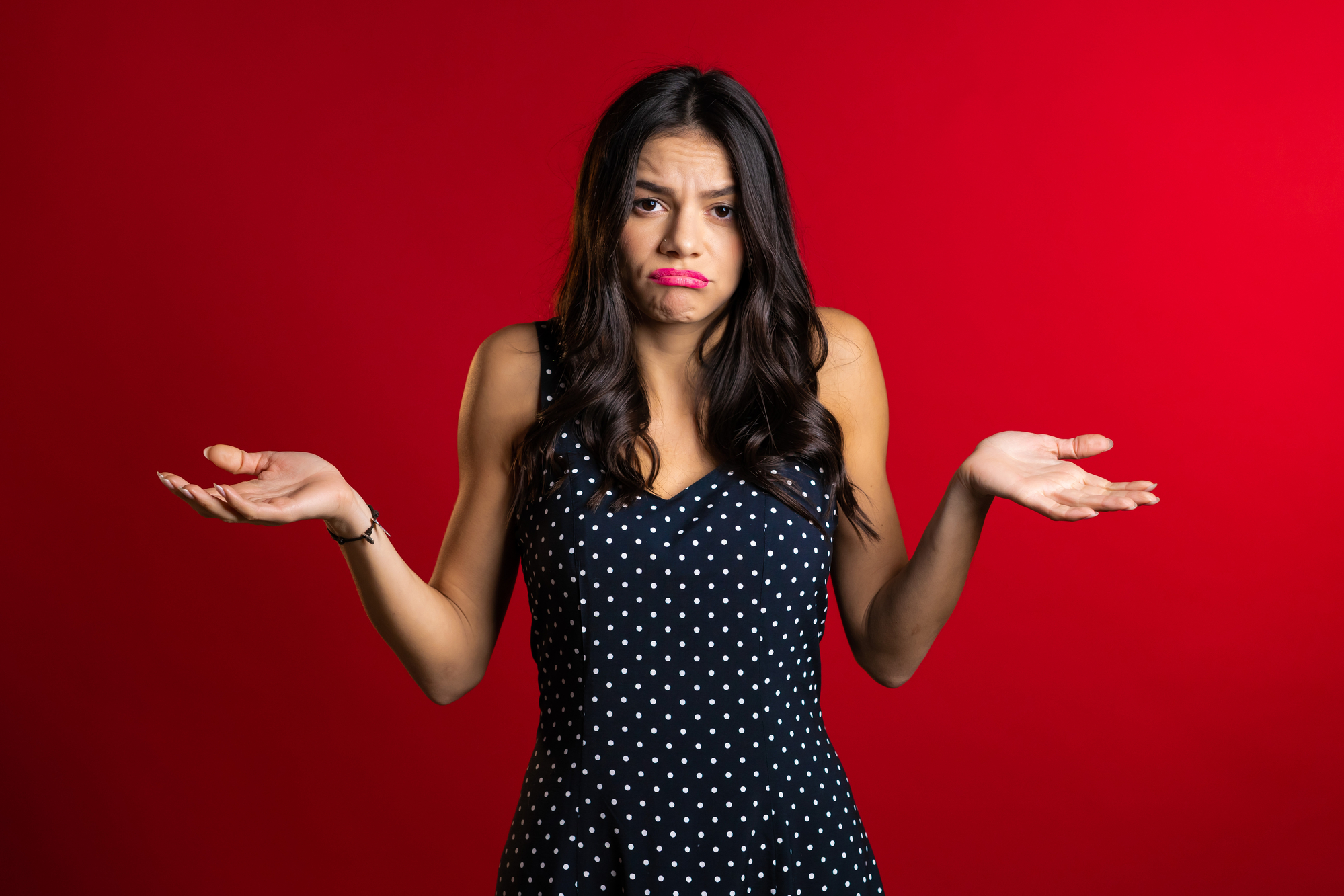 A woman with long dark hair, wearing a black dress with white polka dots, stands against a solid red background. She has a puzzled expression, with raised eyebrows and pursed lips, and she is shrugging her shoulders with her palms facing up.