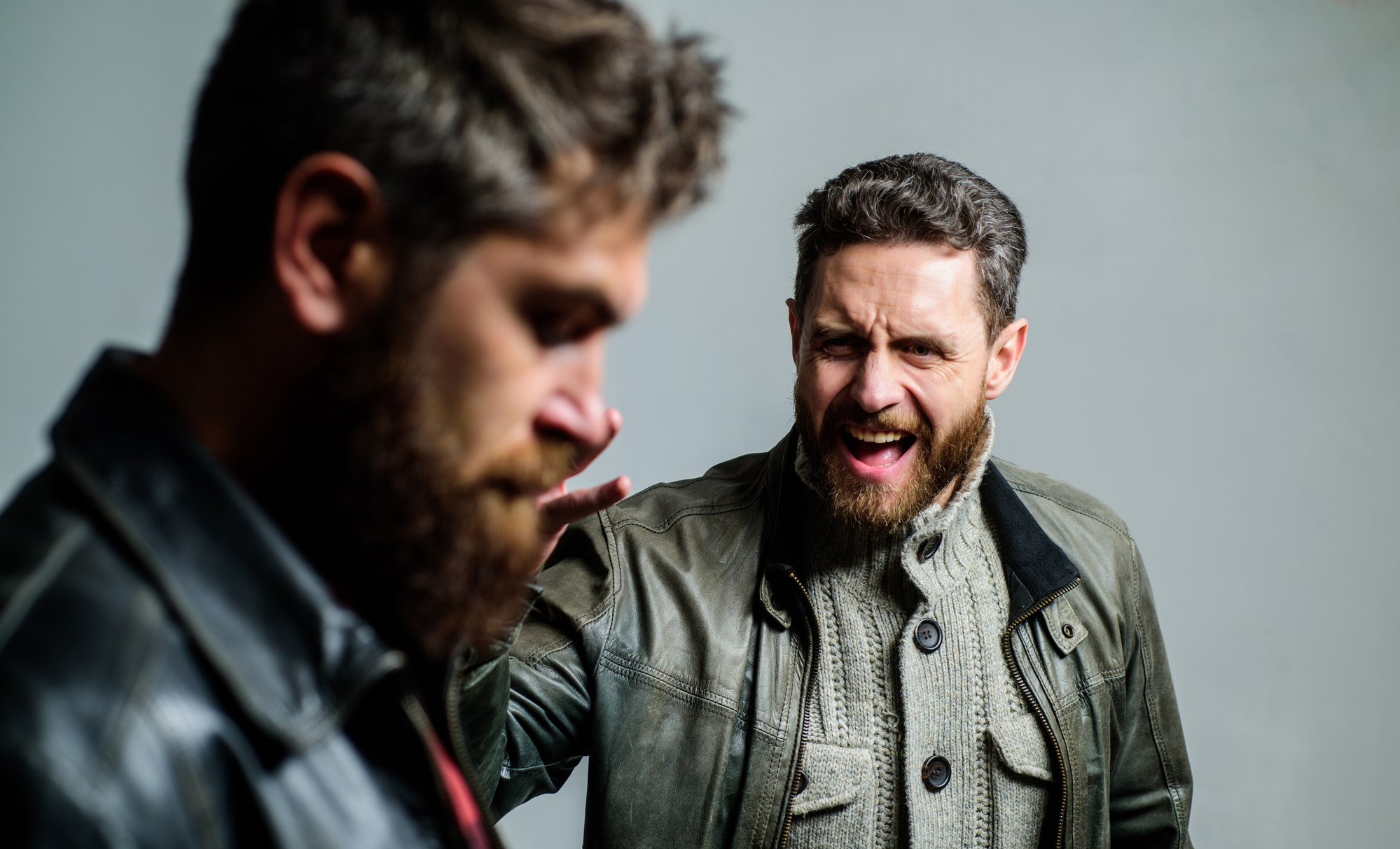 A bearded man in a gray jacket angrily shouts at another bearded man in a leather jacket, who looks downcast while looking away. The background is a plain, light-colored wall.