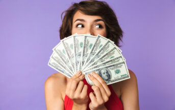 A person with short brown hair and a red sleeveless top holds up a fanned stack of hundred-dollar bills, partially covering their face, while looking to the side. The background is a solid purple color.