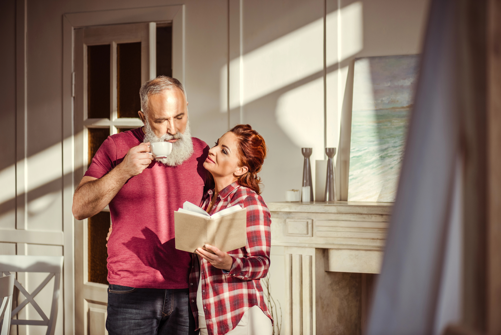 An older man with a beard drinks from a large white cup while a woman with red hair smiles and holds a book. They stand close to each other in a sunlit room with a fireplace and artwork in the background. The atmosphere is warm and cozy.