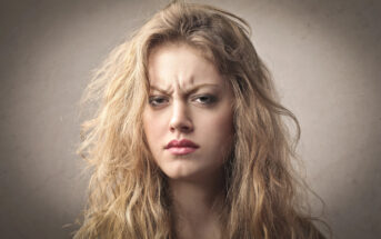 Close-up of a young woman with long, messy blonde hair, looking directly at the camera with a serious and slightly annoyed expression. She has furrowed brows and slightly pursed lips, set against a plain, neutral background.