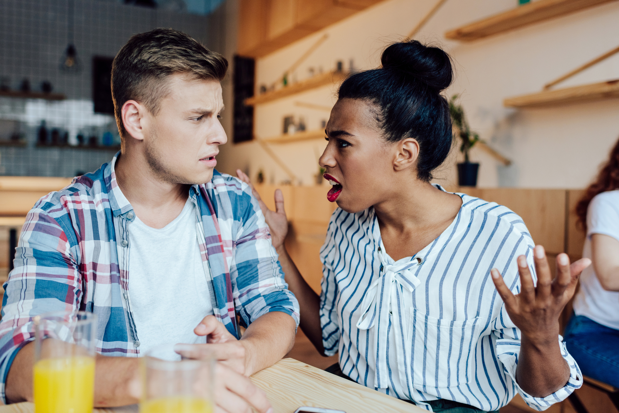 A woman with an expressive face gestures with her hands while talking to a man who looks surprised and confused. They are sitting at a wooden table in a cafe with drinks in front of them. The man wears a plaid shirt, and the woman wears a striped blouse.