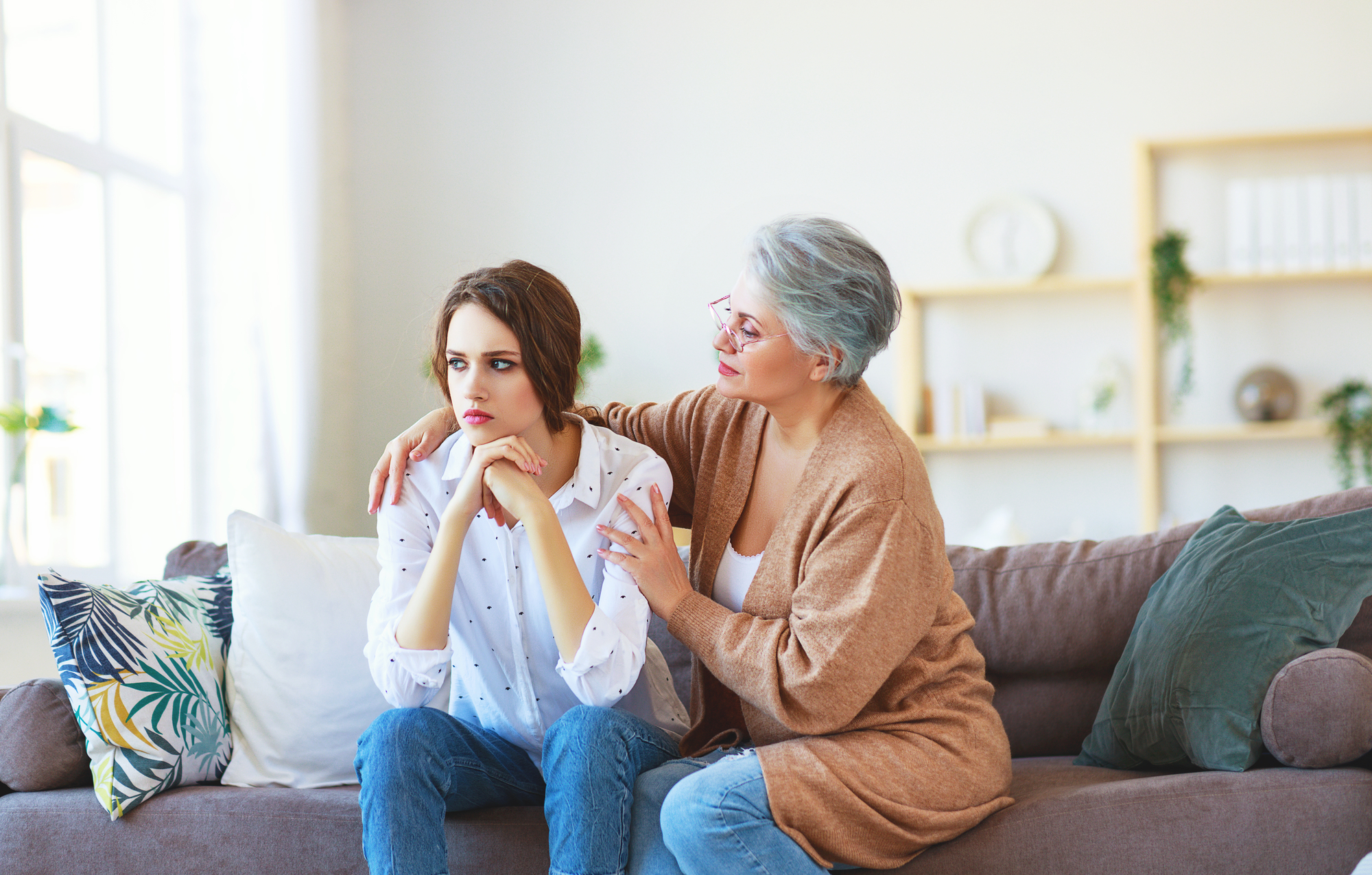 An older woman with gray hair and glasses sits on a sofa, comforting a younger woman with brown hair who looks upset. The older woman has her arm around the younger woman, offering support. They are in a well-lit, cozy living room with plants and shelves in the background.