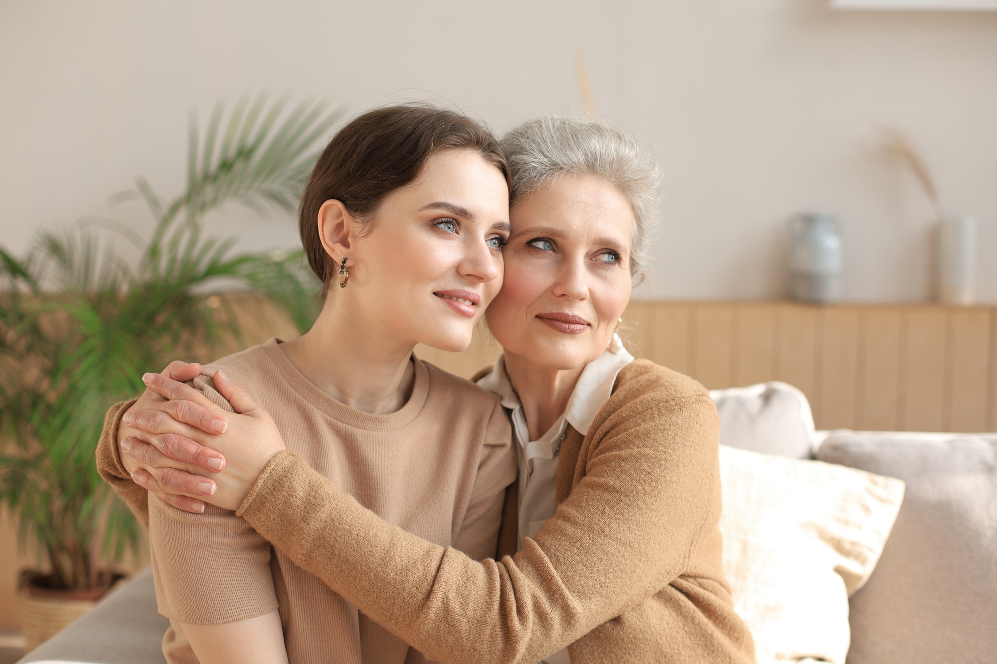 A younger woman and an older woman sit closely together on a light-colored couch. The younger woman has short dark hair and wears a beige top. The older woman has short gray hair and wears a light brown cardigan over a white blouse. They are smiling and embracing.