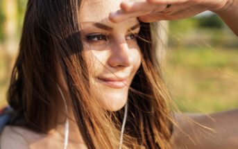 A close-up of a young woman outdoors, shielding her eyes from the sun with one hand while smiling. She has long brown hair with some strands falling across her face, and she is wearing earphones. The background is blurred, showing greenery and sunlight.