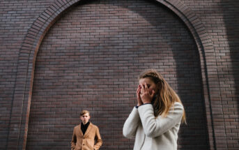 A woman in a white coat stands in the foreground, covering her face with her hands. In the background, a man in a tan coat stands with his hands in his pockets. They are in front of a large, arched brick wall. The scene appears to be emotionally charged.