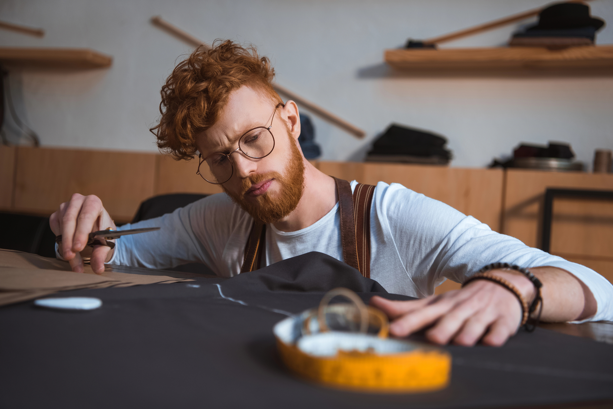 A focused man with curly red hair and glasses cuts fabric on a table. He is wearing a white shirt with brown suspenders and is working in a sewing or tailoring studio, surrounded by shelves holding materials and tools, including scissors and a measuring tape.