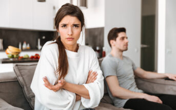 A woman with long hair in a white sweater sits on a couch with her arms crossed and an upset expression. In the background, a man in a gray t-shirt sits on the same couch looking away. They appear to be in a modern living room or kitchen setting.