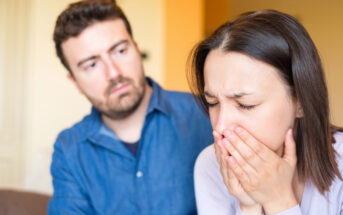 A worried man looks at a distressed woman covering her mouth with her hands, suggesting she is upset or crying. They are indoors, with a blurred background, creating an intimate and concerned ambiance.