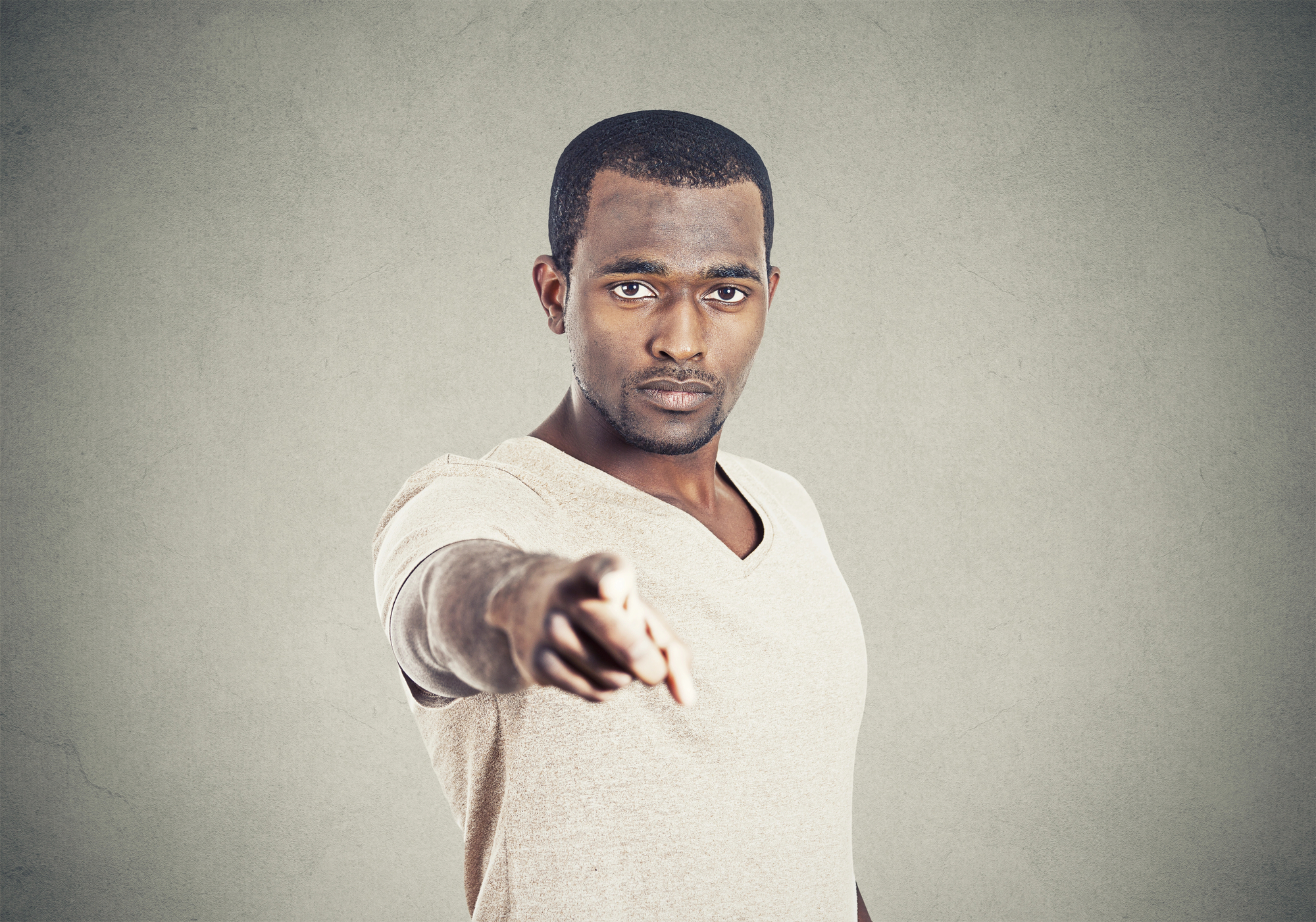 A man with short hair and a trimmed beard is standing against a plain grey background, pointing directly at the camera with a serious expression. He is wearing a light beige t-shirt.