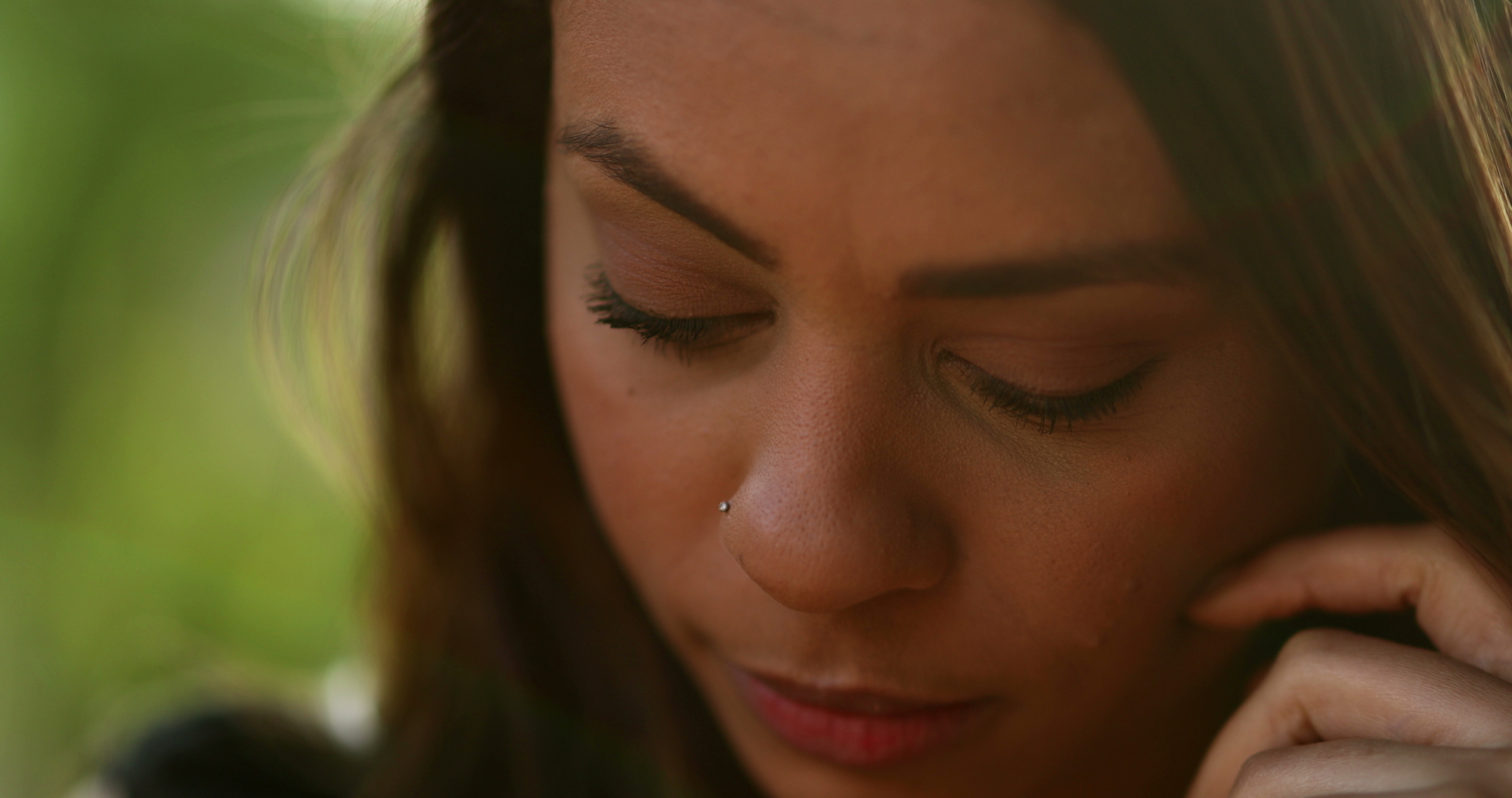 Close-up of a woman with long hair, looking down and touching her face gently. She has a nose stud and appears to be deep in thought or sad. The background is blurred with natural green tones.