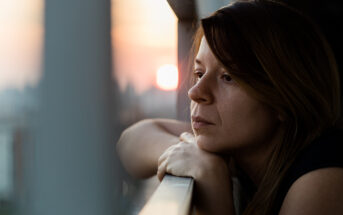 A woman with long brown hair leans on a railing, gazing pensively into the distance during sunset. The sky is tinged with warm pink and orange hues, and the soft light casts gentle shadows on her contemplative face.