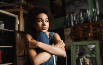 A young woman with natural curly hair sits pensively with her arms wrapped around her knees in a rustic room filled with shelves, bottles, and wooden furniture. She wears a denim outfit and looks thoughtfully into the distance.