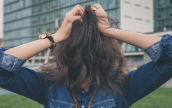 A person stands outdoors in front of a modern building, wearing a denim shirt and bracelets. They have their hands in their long, wavy brown hair, covering their face.