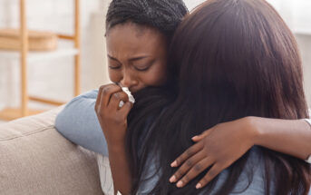 Two women are seen embracing on a couch. One woman is crying and holding a tissue to her nose while being comforted by the other woman. The comforting woman has her arm wrapped around the crying woman, providing support and solace.