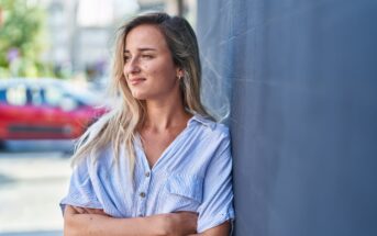 A young woman with long blonde hair and wearing a light blue, striped shirt stands against a dark gray wall. She is smiling slightly and looking to her left. A red car and blurry greenery are visible in the background.