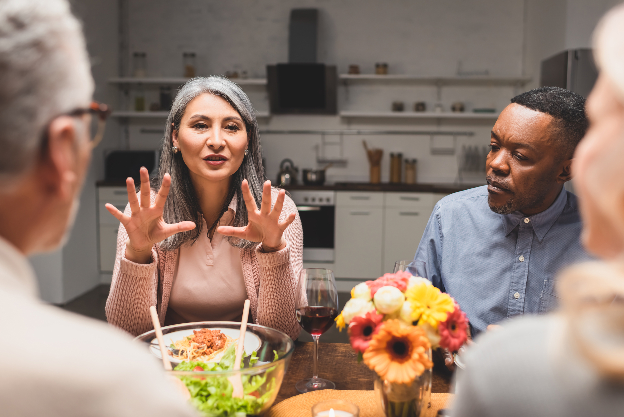 A group of people sit around a dining table engaging in conversation. A woman with gray hair gestures with her hands while speaking. A man in a blue shirt listens attentively. The table has food, including a salad, and a vase of colorful flowers.